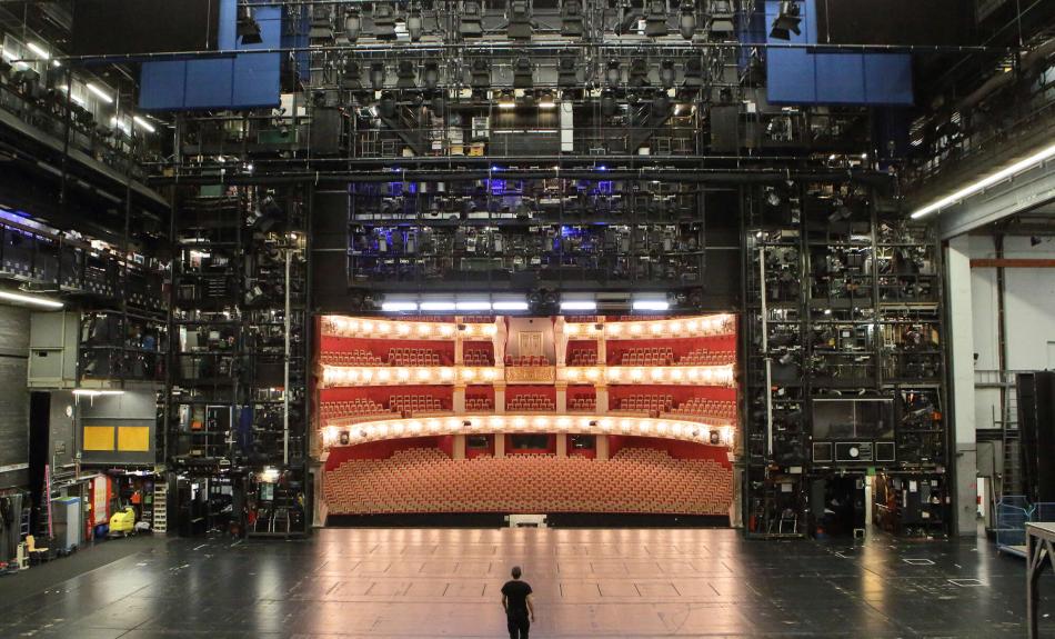 Eine Person steht auf einer Theaterbühne und blickt in den halbrunden Zuschauerraum