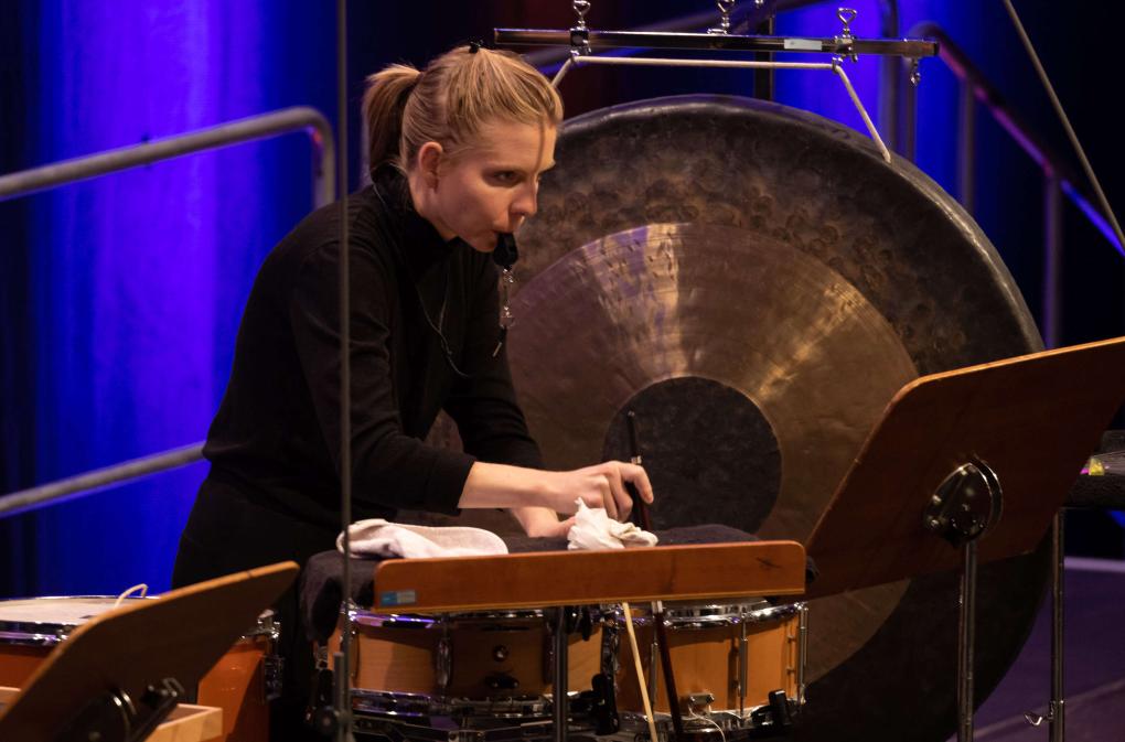 Die Musikerin steht vor einem Gong, hat eine Trillerpfeiffe im Mund blickt konzentriert aufs Notenpult