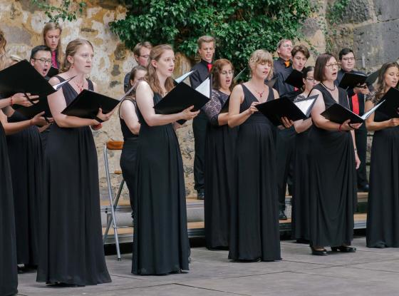 Ein schwarz gekleideter Chor singt im Freien vor einer Mauer aus schwarzen Notenmappen